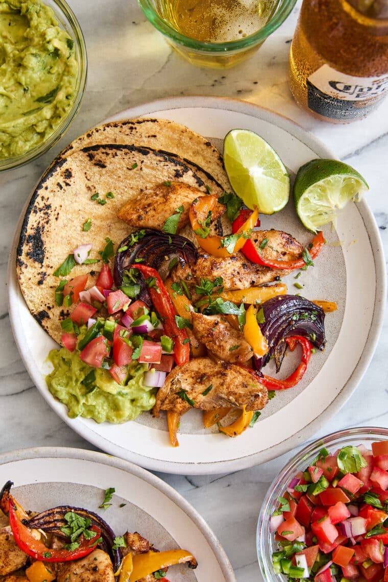 Sheet pan chicken fajitas with sliced chicken, red, yellow, and green bell peppers, red onions, lime wedges, and cilantro on a wooden surface.
