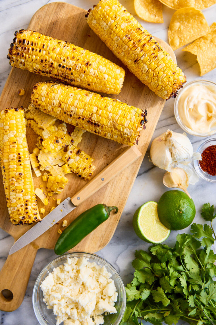 Close-up of creamy Mexican street corn dip with roasted corn, herbs, crumbled cheese, and chili powder in a brown bowl, with tortilla chips nearby.