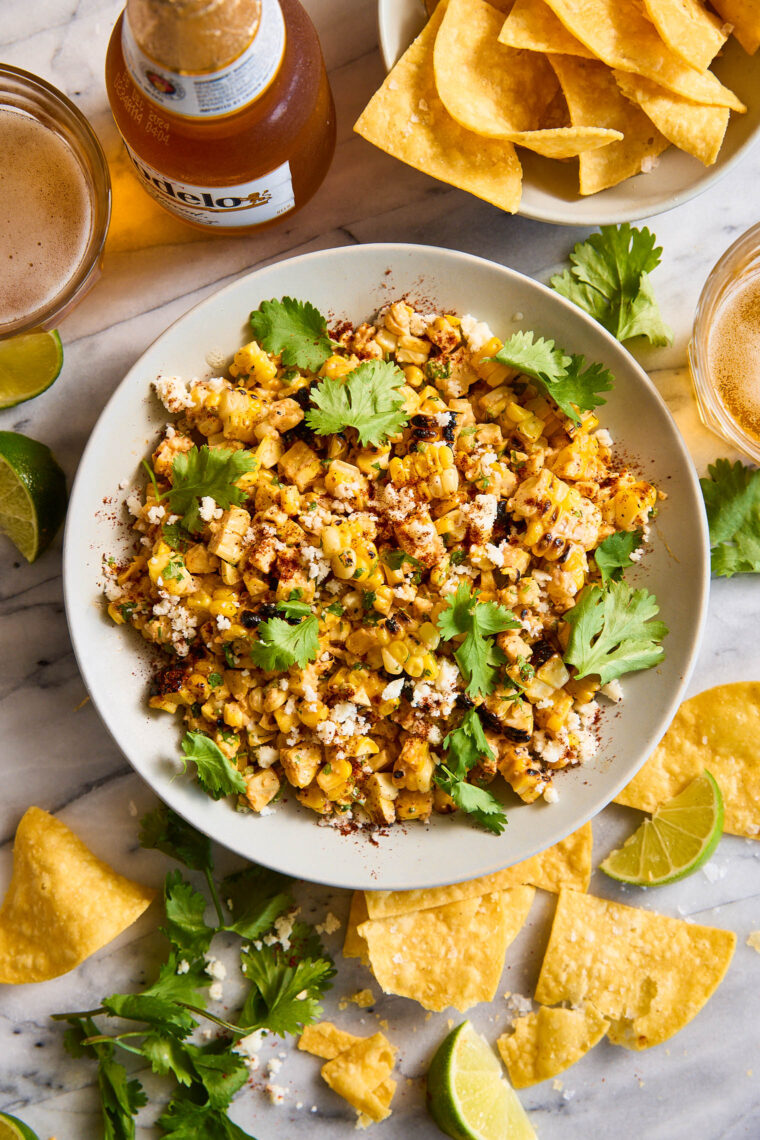 Mexican street corn dip with charred corn, herbs, crumbled cheese, and chili powder in a bowl, served with tortilla chips.