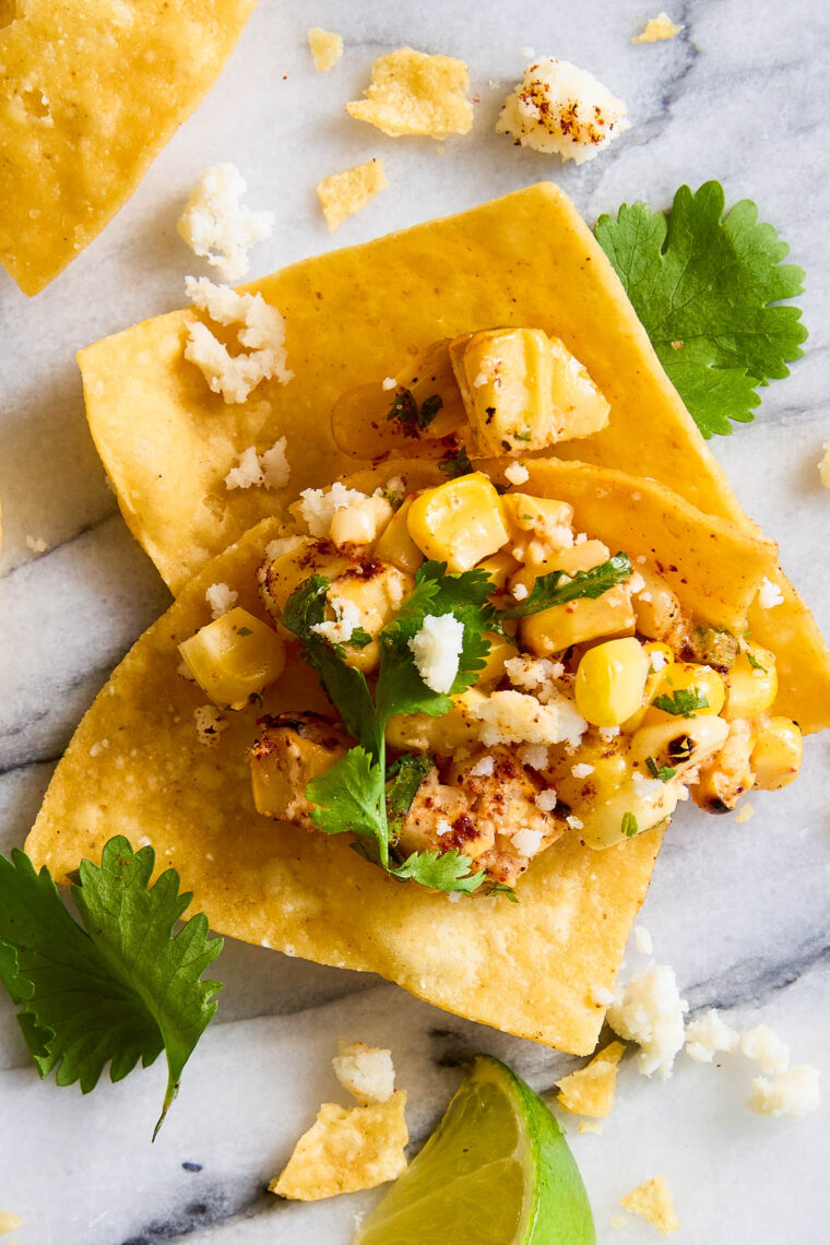 Close-up of tortilla chip lifting creamy Mexican corn dip with roasted corn, herbs, crumbled cheese, and chili powder from a bowl.